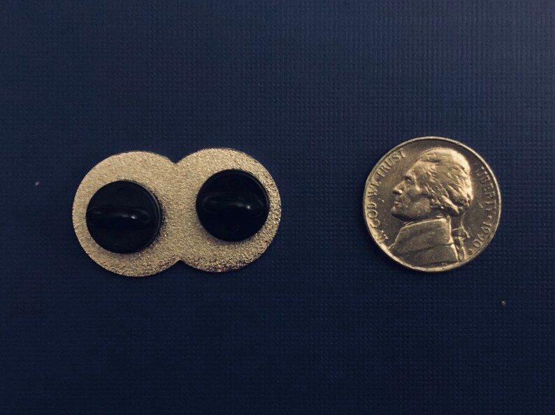 K&ouml;nnte beinhalten: R&uuml;ckansicht eines goldfarbenen Doppelkreis-Pins mit schwarzen Mittelpunkten. Der Pin ist etwa so gro&szlig; wie eine deutsche F&uuml;nf-Cent-M&uuml;nze.