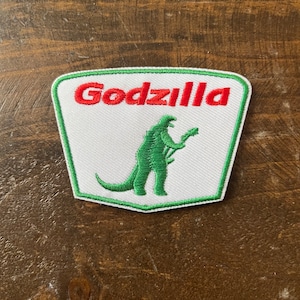 May include: A white, green, and red embroidered patch with the word "Godzilla" in red and a green Godzilla silhouette.