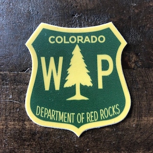 May include: A green and yellow shield-shaped sticker with the text "COLORADO WAP DEPARTMENT OF RED ROCKS". A stylized pine tree is in the center of the sticker.