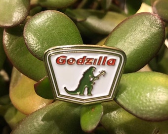 May include: A white enamel pin with a red and white "Godzilla" logo and a green Godzilla silhouette holding a guitar.