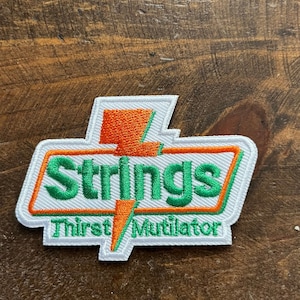 May include: Embroidered patch with a white border, featuring the word "Strings" in green, and "Thirst Mutilator" below. An orange lightning bolt design is above the word "Strings". The patch is on a wooden surface.