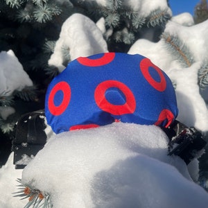 May include: A blue ski goggle cover with a red donut pattern. The goggle cover is partially covered in snow.