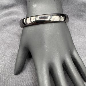 May include: A shiny, silver-colored bangle bracelet. The bracelet is a solid band with a smooth, reflective surface. The bracelet is displayed on a mannequin hand.