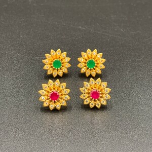 Può includere: Un paio di orecchini dorati a forma di fiore con una pietra preziosa verde e rossa al centro.
