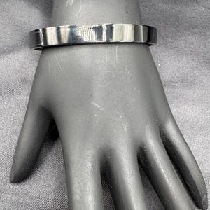 May include: A silver-colored, flat-top bracelet. The bracelet is a solid band with a smooth, reflective surface. The bracelet is shown on a mannequin hand.