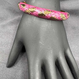 May include: A pink bangle bracelet with gold-colored palm tree designs. The bracelet is on a black hand. The bracelet has a gold-colored clasp.