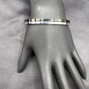 May include: A silver-colored, rectangular bracelet with a flat top and bottom. The bracelet is worn on a mannequin hand, showcasing its design. The bracelet is a simple, elegant piece of jewelry.