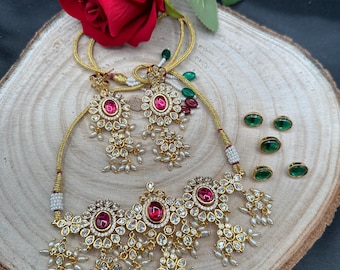 May include: A gold-toned jewelry set featuring a necklace and earrings, adorned with red gemstones and clear crystals. The necklace and earrings have a floral design with dangling pearls. Also included are three pairs of green stud earrings. A red rose rests in the background.