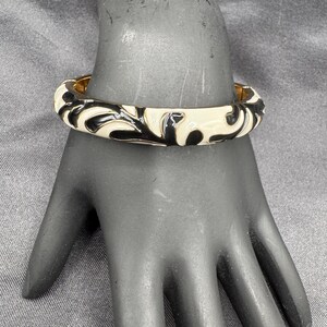 May include: A black and white enamel bangle bracelet with a gold-tone metal band. The bracelet features a repeating pattern of black swirls on a cream-colored background. The bracelet is displayed on a black hand.