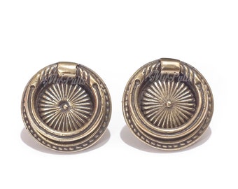 May include: Two antique brass cabinet knobs with a circular design and a raised ring around the edge. The knobs have a sunburst pattern in the center.