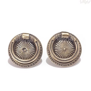 May include: Two antique brass cabinet knobs with a circular design and a raised ring around the edge. The knobs have a sunburst pattern in the center.