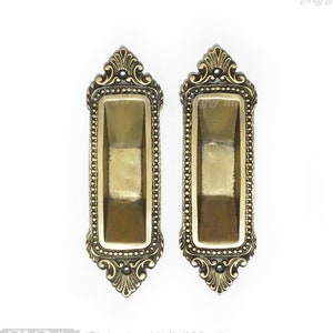 May include: Two ornate gold-toned cabinet door pulls with a decorative border and a rectangular inset.