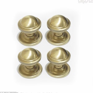 Puede incluir: Cuatro pomos de armario dorados con una parte superior redonda ligeramente abovedada y una base estriada. Los pomos están dispuestos en una cuadrícula de 2x2.