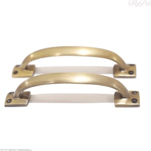 Op de afbeelding: Twee antieke messing kastgrepen met een gebogen ontwerp en vierkante bases. De grepen zijn horizontaal gerangschikt, de ene boven de andere.