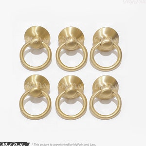 Könnte beinhalten: Sechs goldfarbene Metall-Ringgriffe mit runder Basis. Die Griffe haben eine glatte, polierte Oberfläche.