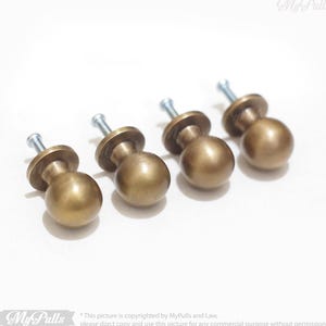 May include: Four antique brass cabinet knobs with a round shape and a screw on the back. The knobs are arranged in a row on a white surface.