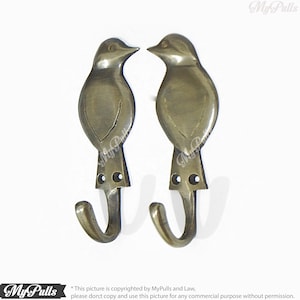Könnte beinhalten: Zwei antike Messing-Vogelhaken mit einem detaillierten Design. Die Haken sind in Form von Vögeln mit ausgebreiteten Flügeln gestaltet. Die Haken sind an einem weißen Hintergrund befestigt.