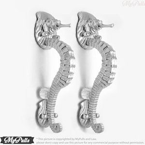 Op de afbeelding: Twee zilverkleurige deurgrepen in de vorm van een zeepaardje. De grepen zijn gedetailleerd met een textuur, schubbenontwerp en hebben een gebogen vorm.