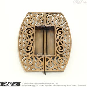May include: A pair of ornate brass door pulls with a decorative, intricate, cutout design. The pulls are oval shaped and have a rectangular opening in the centre.