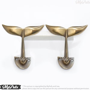 May include: Two antique brass whale tail hooks. The hooks are mounted on a white surface. The hooks are shaped like a whale tail and have a decorative design.