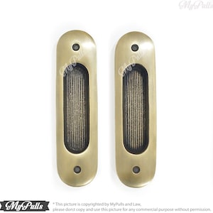 Könnte beinhalten: Zwei antike, messingfarbene Einlassgriffe für Schranktüren oder Schubladen. Jeder Griff hat eine ovale Form mit gerillter Mitte und zwei Schraubenlöchern. Die Griffe sind vor weißem Hintergrund abgebildet. Das My Pulls-Logo befindet sich unten links.