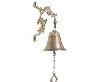 May include: A brass bell with an ornate design hangs from a decorative wall mount. The bell has a chain attached to the bottom.