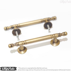 Pode incluir: Duas puxadores de arm&aacute;rio de lat&atilde;o antigo com uma barra cil&iacute;ndrica e detalhes decorativos nas extremidades. Cada puxador inclui uma base redonda e ferragens de montagem. Os puxadores s&atilde;o de cor lat&atilde;o envelhecido e est&atilde;o sobre um fundo branco.