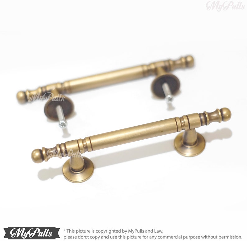 Pode incluir: Duas puxadores de arm&aacute;rio de lat&atilde;o antigo com uma barra cil&iacute;ndrica e detalhes decorativos nas extremidades. Cada puxador inclui uma base redonda e ferragens de montagem. Os puxadores s&atilde;o de cor lat&atilde;o envelhecido e est&atilde;o sobre um fundo branco.