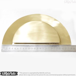 May include: A pair of brass half-circle cabinet pulls, one slightly smaller than the other, with a brushed finish. The pulls are shown against a white background with a ruler for scale.