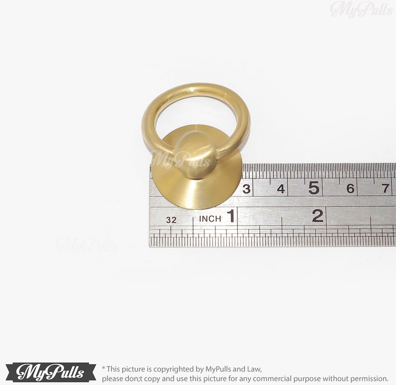 Puede incluir: Una manija de anillo de tracci&oacute;n de lat&oacute;n con una base redonda, que mide 2,5 cm de di&aacute;metro. La manija se muestra junto a una regla para referencia de tama&ntilde;o.