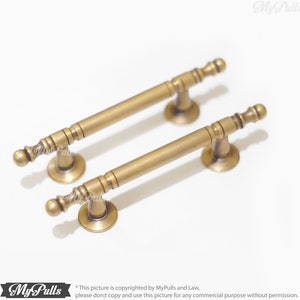 Pode incluir: Duas puxadores de arm&aacute;rio de lat&atilde;o antigo. Cada puxador tem uma barra cil&iacute;ndrica com detalhes decorativos e extremidades arredondadas. Os puxadores s&atilde;o montados em bases circulares. O hardware &eacute; exibido em um fundo branco.