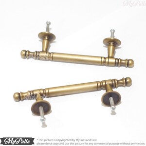 Pode incluir: Duas puxadores de arm&aacute;rio de lat&atilde;o antigo. Cada puxador tem uma al&ccedil;a cil&iacute;ndrica com detalhes decorativos e extremidades arredondadas. Os puxadores s&atilde;o fixados a uma base redonda com um parafuso para montagem. A imagem est&aacute; em um fundo branco.