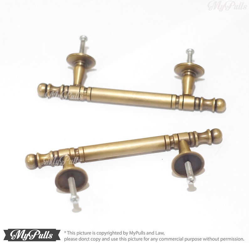 Pode incluir: Duas puxadores de arm&aacute;rio de lat&atilde;o antigo. Cada puxador tem uma al&ccedil;a cil&iacute;ndrica com detalhes decorativos e extremidades arredondadas. Os puxadores s&atilde;o fixados a uma base redonda com um parafuso para montagem. A imagem est&aacute; em um fundo branco.