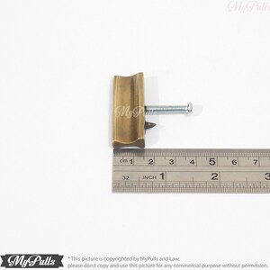 May include: A brass cabinet pull with a curved design and a screw. The pull is 2.5 cm long.