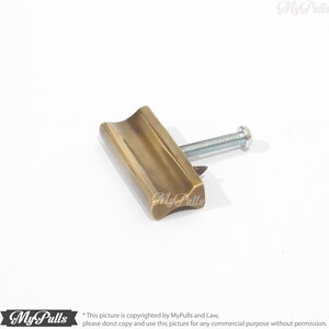 May include: A brass cabinet pull with a rectangular shape and a slightly curved top. The pull has a single screw hole and a brushed finish.