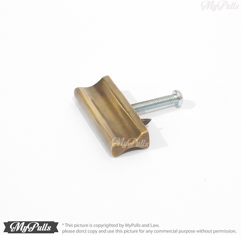May include: A brass cabinet pull with a rectangular shape and a slightly curved top. The pull has a single screw hole and a brushed finish.