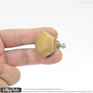 Puede incluir: Un pomo de armario hexagonal dorado con acabado cepillado. El pomo se sostiene en una mano.