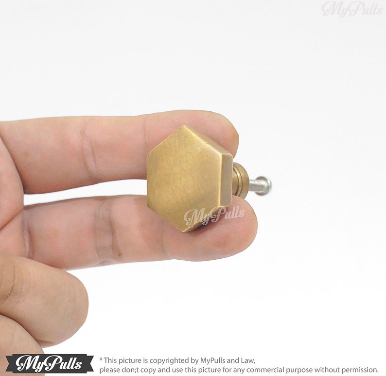 Puede incluir: Un pomo de armario hexagonal dorado con acabado cepillado. El pomo se sostiene en una mano.