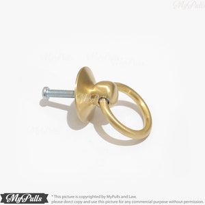 Puede incluir: Tirador de armario de anillo dorado con base redonda y tornillo. El tirador es de metal y tiene un acabado liso.