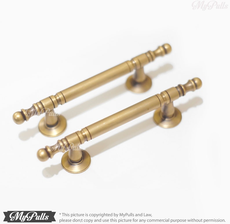 Pode incluir: Duas puxadores de arm&aacute;rio de lat&atilde;o antigo. Cada puxador tem uma barra cil&iacute;ndrica com detalhes decorativos e extremidades arredondadas. Os puxadores s&atilde;o montados em bases circulares. O hardware &eacute; exibido em um fundo branco.