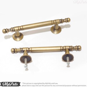 Pode incluir: Duas puxadores de arm&aacute;rio de lat&atilde;o antigo. Cada puxador tem uma barra cil&iacute;ndrica com detalhes decorativos e extremidades arredondadas. Os puxadores s&atilde;o montados em uma base circular com um parafuso para instala&ccedil;&atilde;o. Fundo branco.