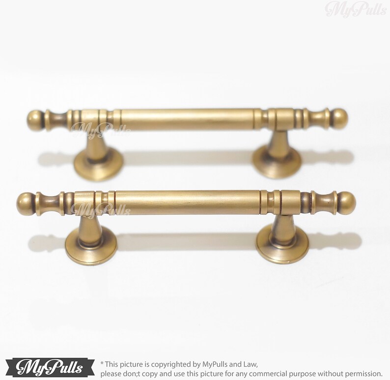 Pode incluir: Duas puxadores de arm&aacute;rio de lat&atilde;o antigo. Cada puxador tem uma barra cil&iacute;ndrica com extremidades decorativas e uma base alargada. Os puxadores s&atilde;o exibidos em um fundo branco. O logotipo My Pulls est&aacute; no canto inferior esquerdo.