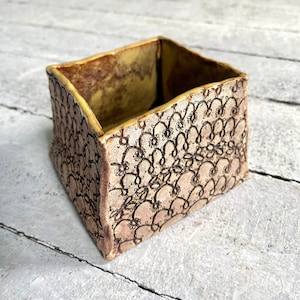 May include: A square ceramic planter with a brown and yellow glaze. The planter has a decorative lace pattern carved into the surface.
