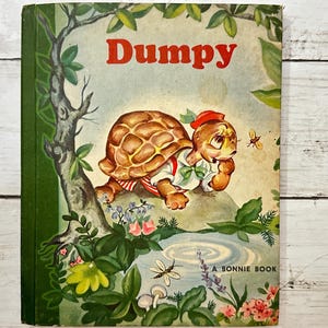 Puede incluir: Una portada de libro infantil vintage con una tortuga de dibujos animados llamada "Dumpy" que lleva un sombrero rojo y una camisa verde. La tortuga camina por un bosque verde con flores y un estanque. El título del libro es "Dumpy" y el libro es un "Bonnie Book".