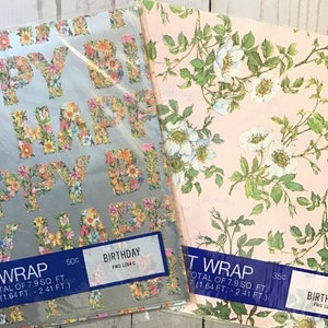 May include: Two rolls of floral patterned gift wrap. The first roll is silver with a floral pattern spelling out "Happy Birthday". The second roll is pink with a white floral pattern. Both rolls have blue labels with the text "Wrap" and the price.