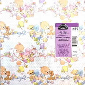 Op de afbeelding: Een witte cadeaupapierrol met een babythema. Het papier heeft een herhalend patroon van schattige babyfiguren, speelgoed en ballonnen. De tekst "Expressions Hallmark Gift Wrap" staat op het papier gedrukt, samen met de prijs en de afmetingen.