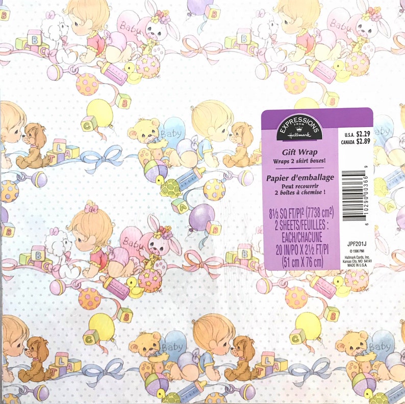 Op de afbeelding: Een witte cadeaupapierrol met een babythema. Het papier heeft een herhalend patroon van schattige babyfiguren, speelgoed en ballonnen. De tekst "Expressions Hallmark Gift Wrap" staat op het papier gedrukt, samen met de prijs en de afmetingen.
