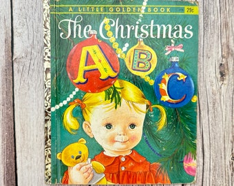 Pode incluir: Um livro vintage Little Golden Book intitulado "The Christmas ABC" com uma capa verde que apresenta uma menina com rabo de cavalo loiro, um urso de pelúcia e enfeites de Natal com as letras A, B e C.