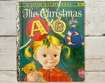 May include: A vintage Little Golden Book titled "The Christmas ABC" with a green cover featuring a girl with blonde hair and a teddy bear. The girl is wearing a red dress and is surrounded by Christmas ornaments shaped like the letters A, B, and C.