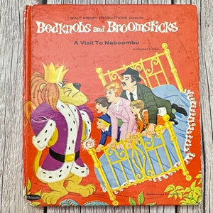 May include: A vintage children's book cover featuring the characters from the Disney movie Bedknobs and Broomsticks. The cover is red with a yellow bed and a lion in a purple robe. The title of the book is "Bedknobs and Broomsticks: A Visit To Naboombu".